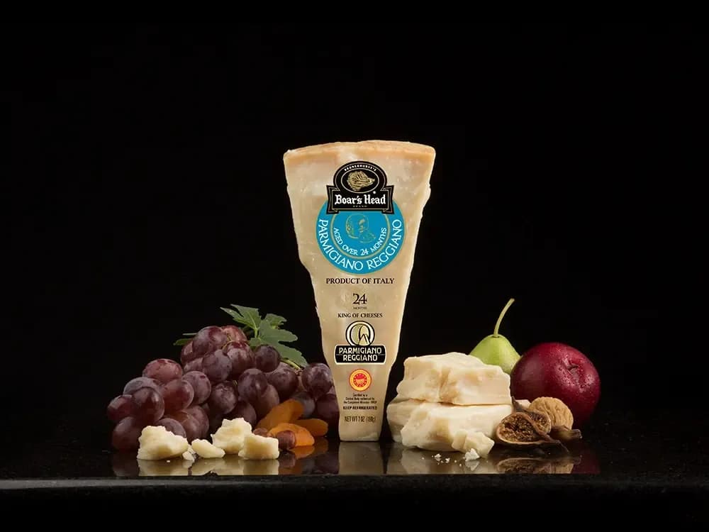 The Boar's Head® Parmigiano Reggiano Cheese sits on a counter top, surrounded by grapes, dried apricots, figs, a pear, a red apple, and some unwrapped and crumbled product, with a black background.