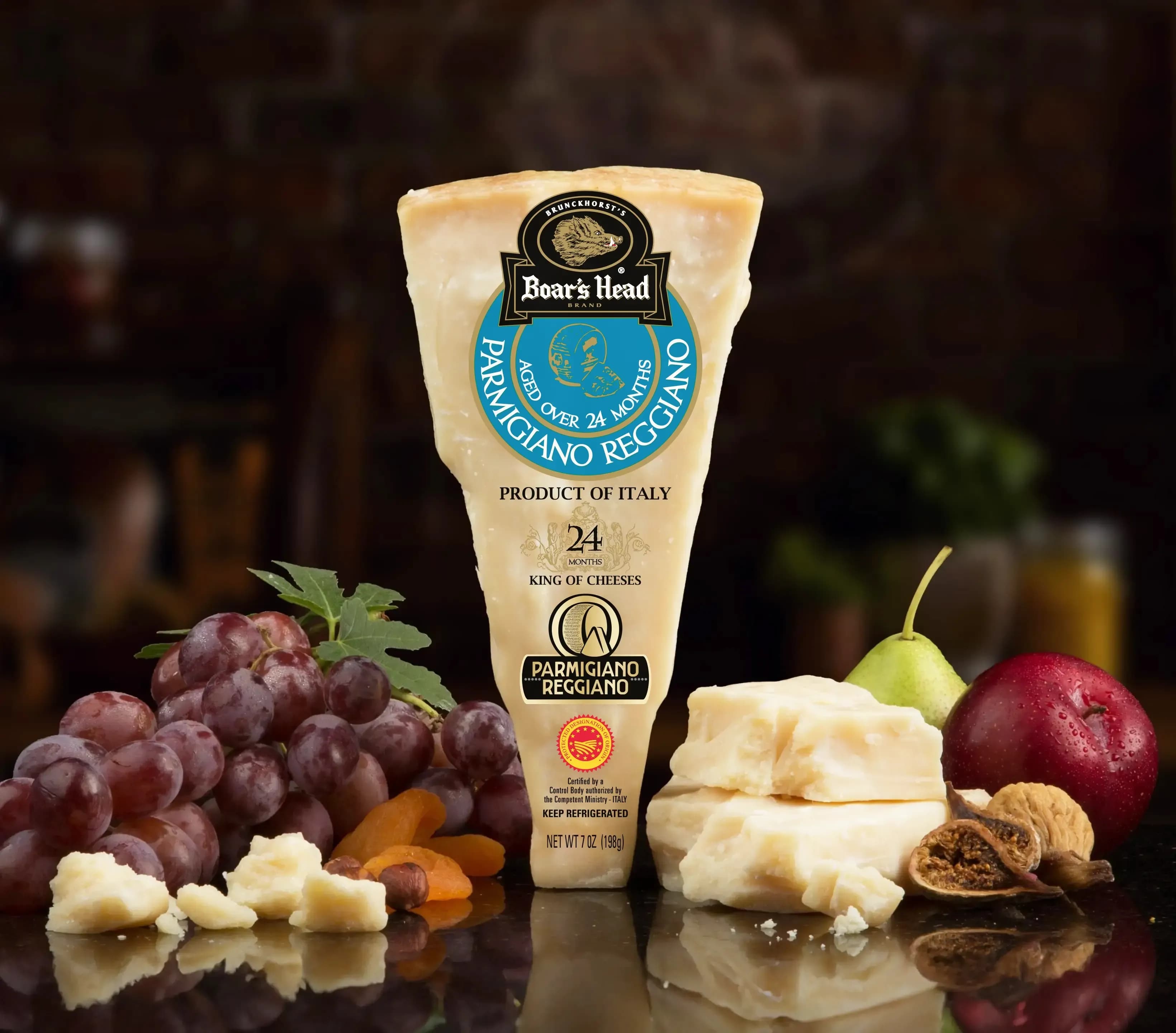 The Boar's Head® Parmigiano Reggiano Cheese sits on a counter top, surrounded by grapes, dried apricots, figs, a pear, a red apple, and some unwrapped and crumbled product, all in front of blurred deli scene in the background.