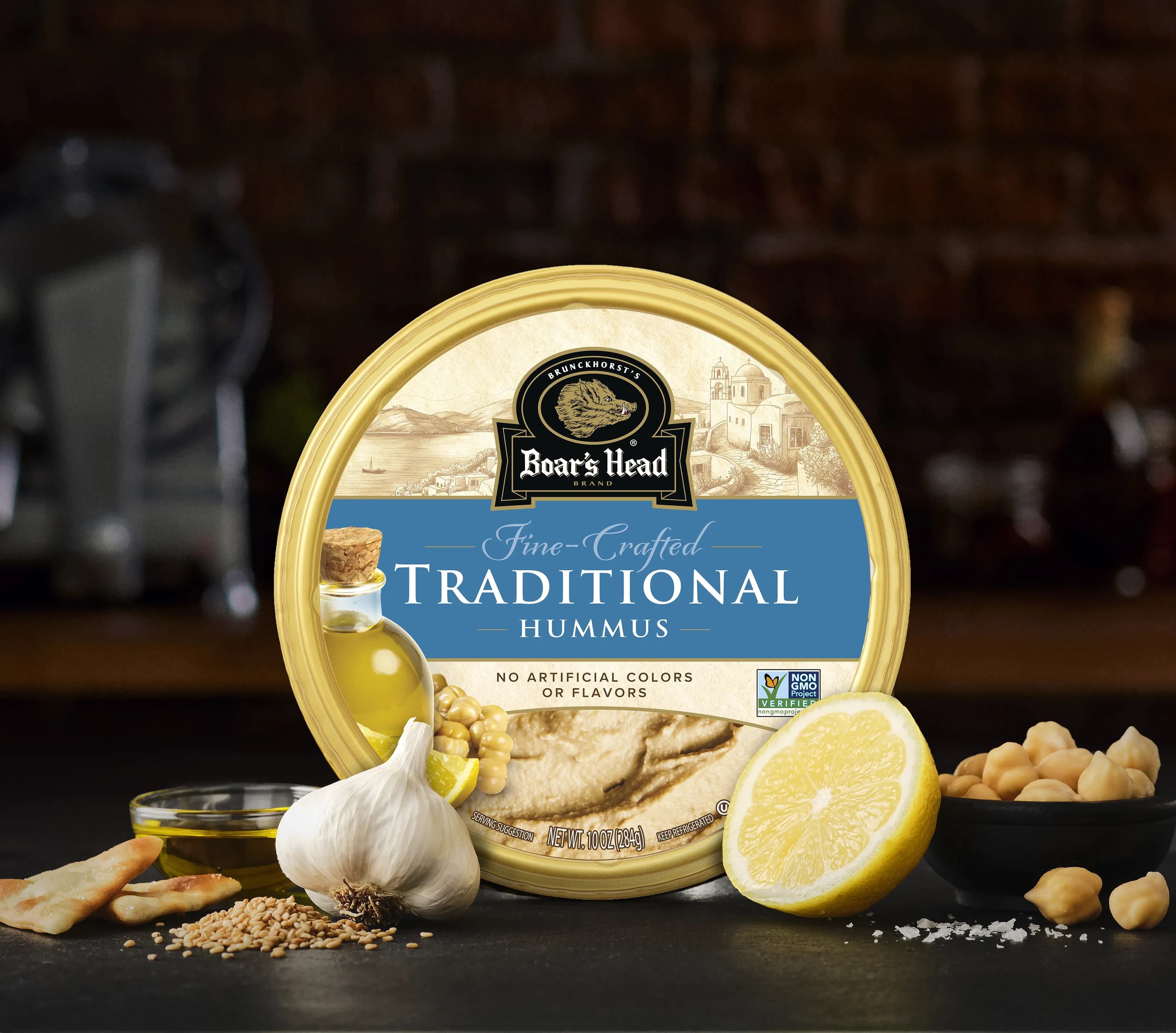 The Boar's Head® Traditional Hummus sits on a counter top, surrounded by garlic, chickpeas, sesame seeds, and pita chips, all in front of blurred deli scene in the background.