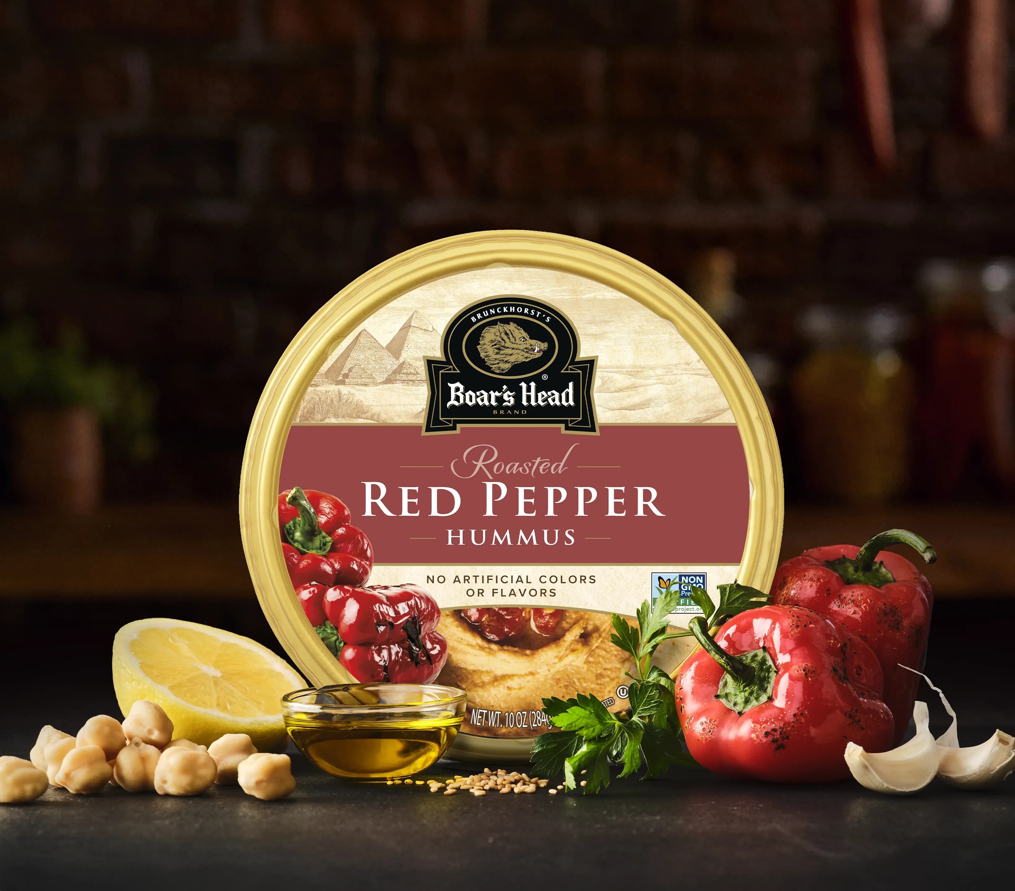 The Boar's Head® Red Pepper Hummus sits on a counter top, surrounded by chickpeas, roasted red peppers, olive oil, and a slice of lemon, all in front of blurred deli scene in the background.