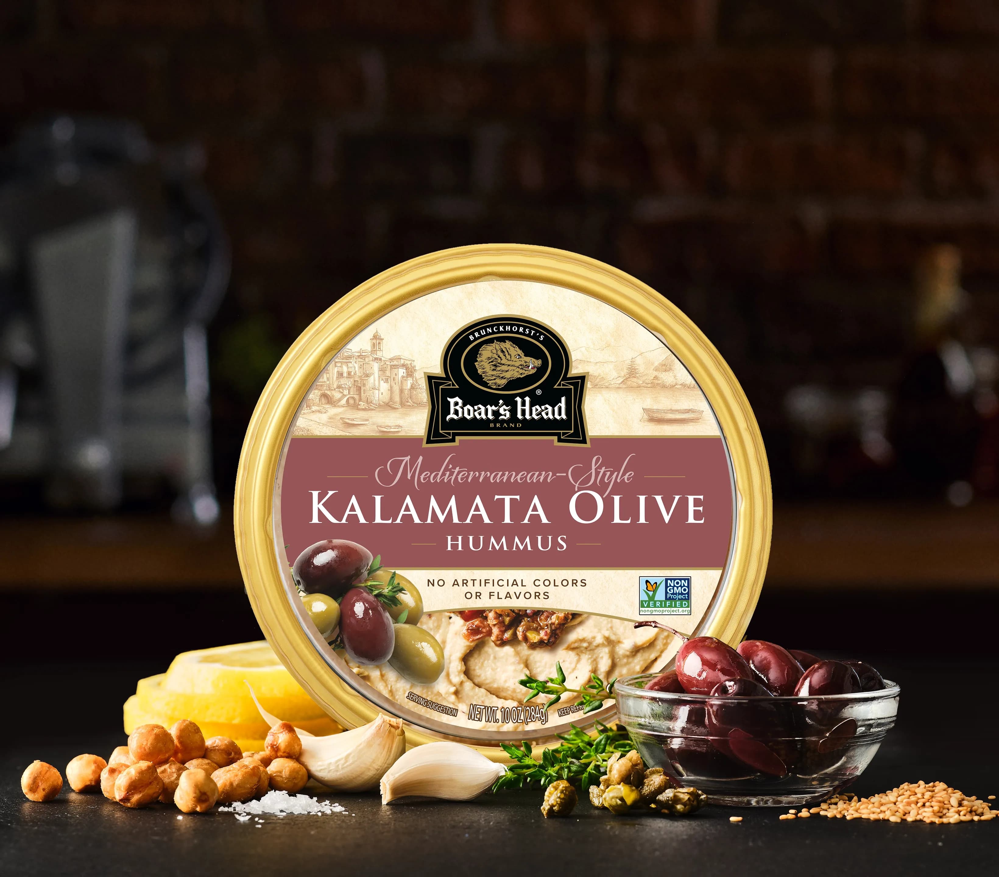 The Boar's Head® Kalamata Olive Hummus sits on a counter top, surrounded by garlic cloves, chickpeas, kalamata olives, and slices of lemon, all in front of blurred deli scene in the background.