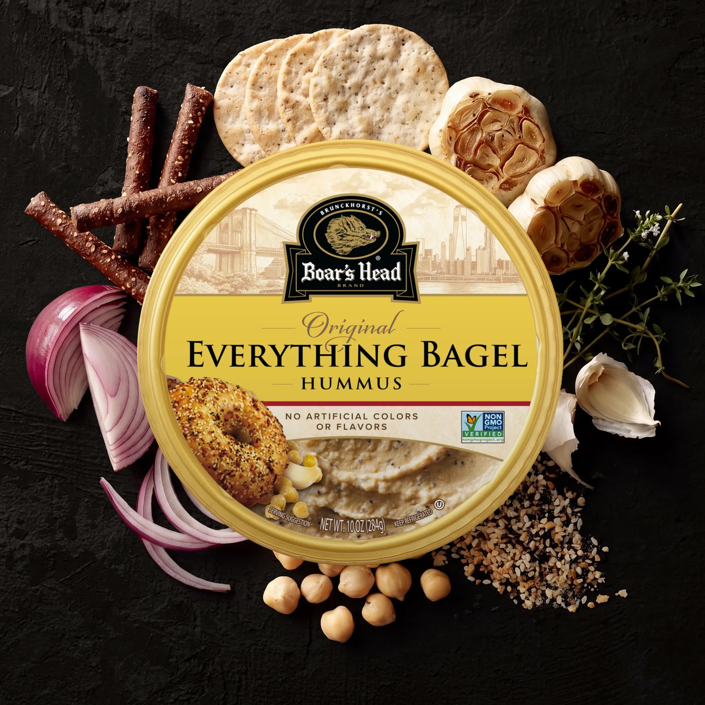 An aerial view of the Boar's Head® Everything Bagel Hummus that sits on a graphite colored background, surrounded by roasted garlic, everything bagel seasoning, red onions, chips, and green garnishes.