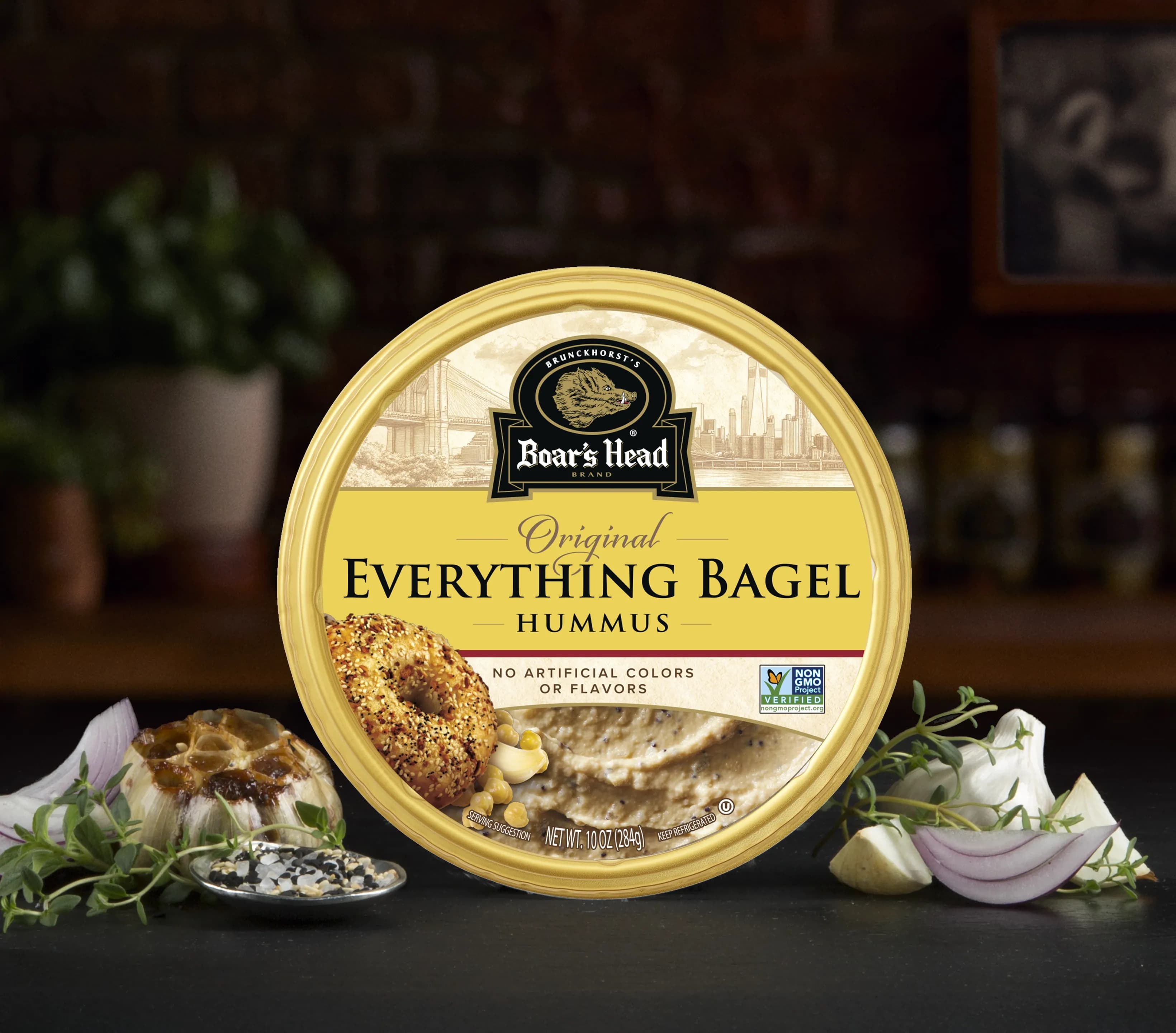 The Boar's Head® Everything Bagel Hummus sits on a counter top, surrounded by roasted garlic, everything bagel seasoning, red onions, chips, and green garnishes, all in front of blurred deli scene in the background.