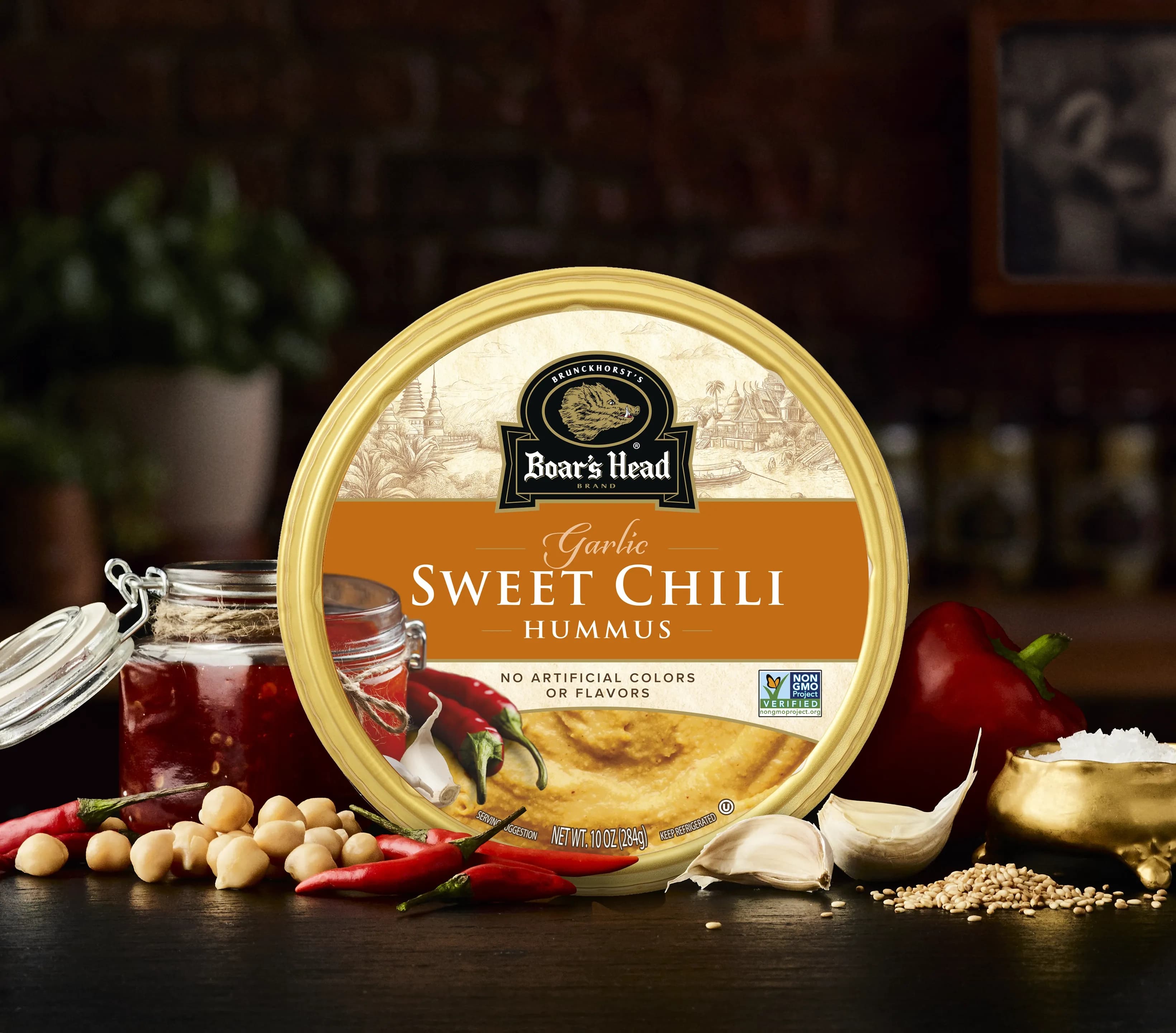 The Boar's Head® Sweet Chili Hummus sits on a counter top, surrounded by sweet chili sauce, garlic, peppers, chickpeas, sesame seeds, and salt, all in front of blurred deli scene in the background.
