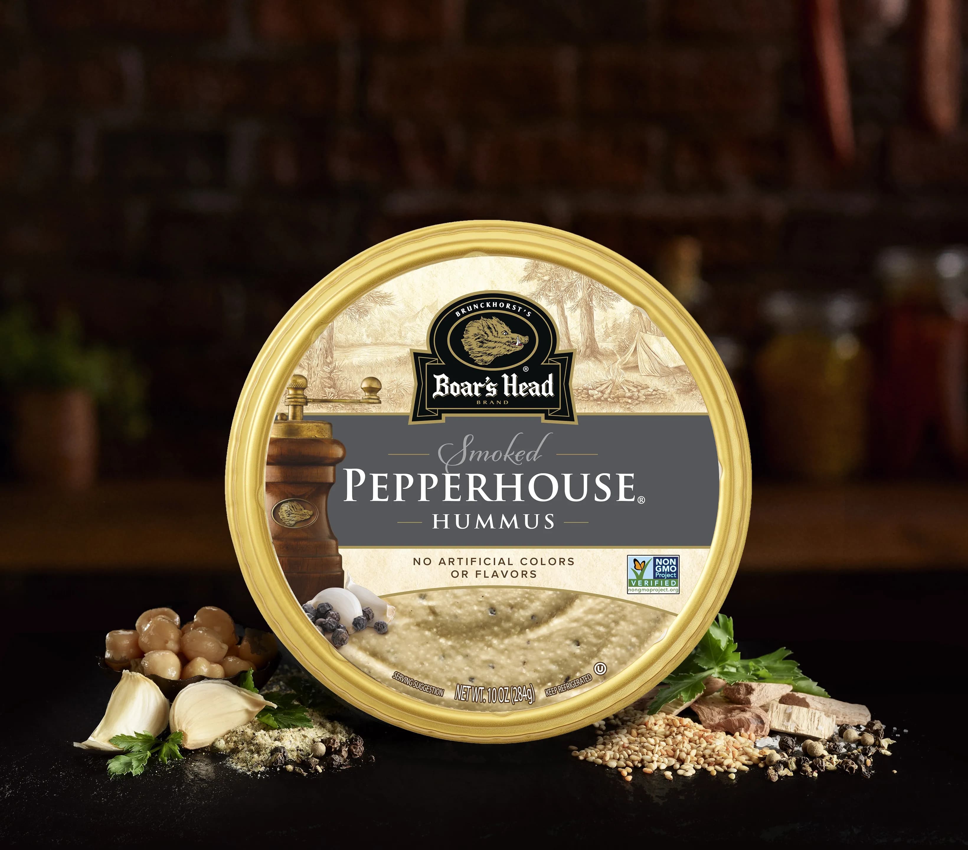 The Boar's Head® PepperHouse® Hummus sits on a counter top, surrounded by red peppers, lemon, chickpeas, and olive oil, all in front of blurred deli scene in the background.