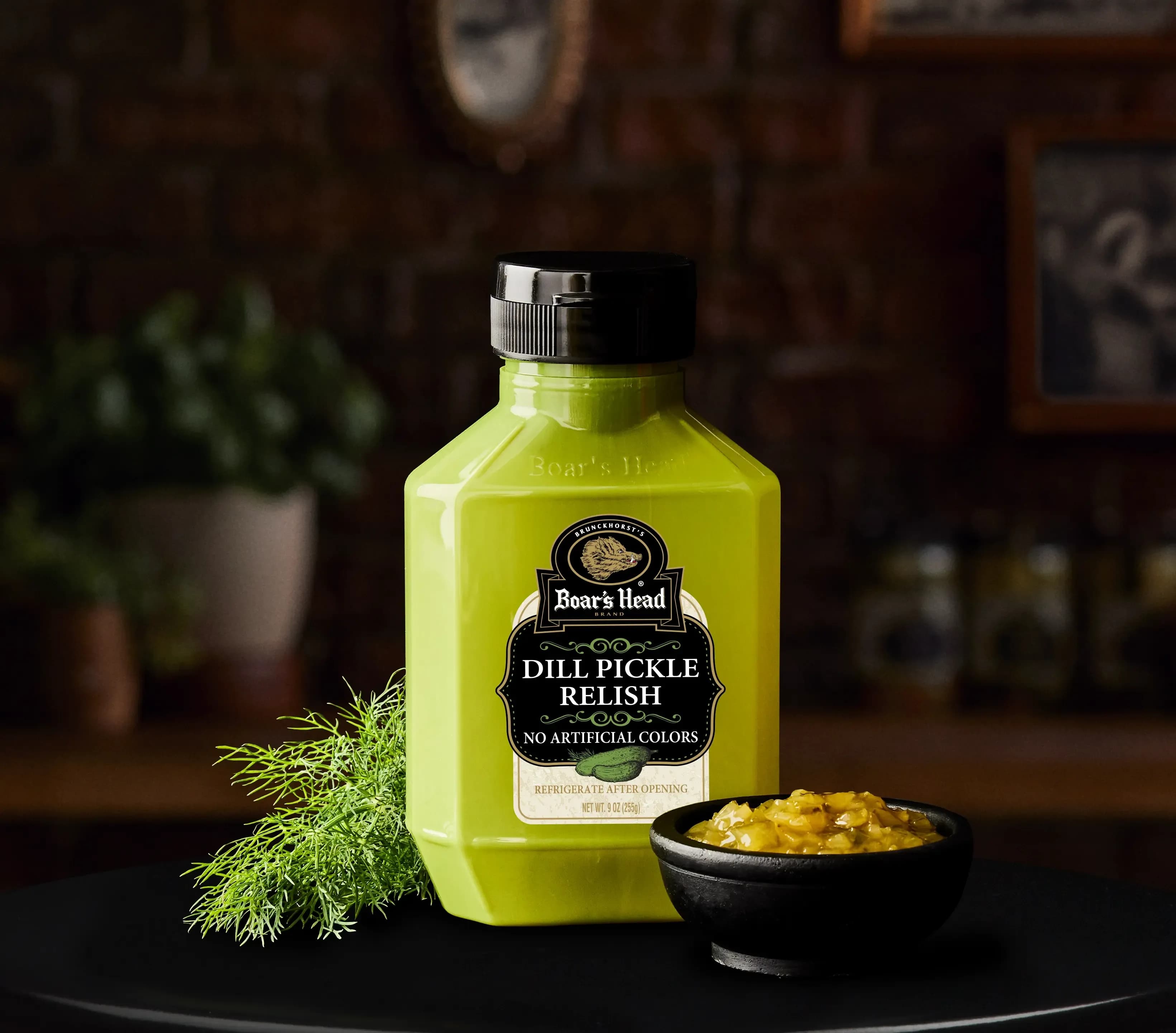 A container of Boar's Head® Dill Pickle Relish sits on a counter top, surrounded by a small bowl of the relish and some dill greenery, all sitting in front of blurred deli scene in the background.