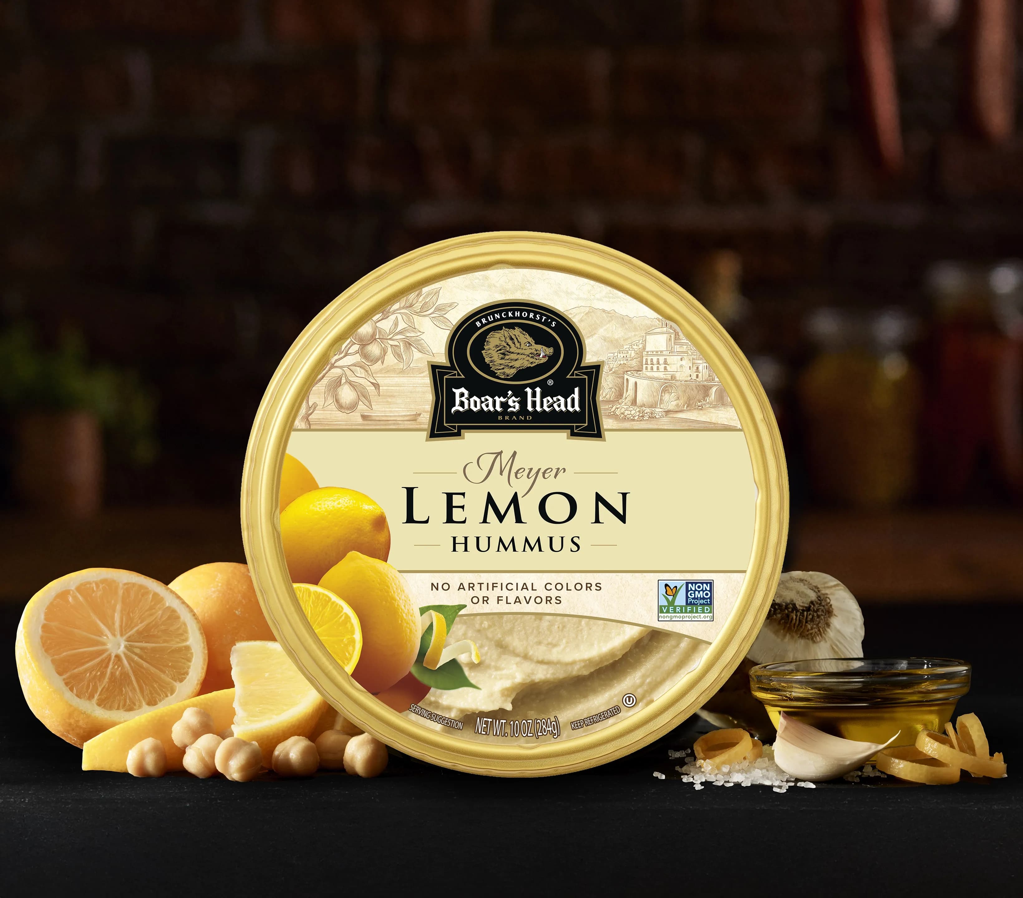 The Boar's Head® Lemon Hummus sits on a counter top, surrounded by slices of lemon, chickpeas, garlic, chips, and olive oil, all in front of blurred deli scene in the background.