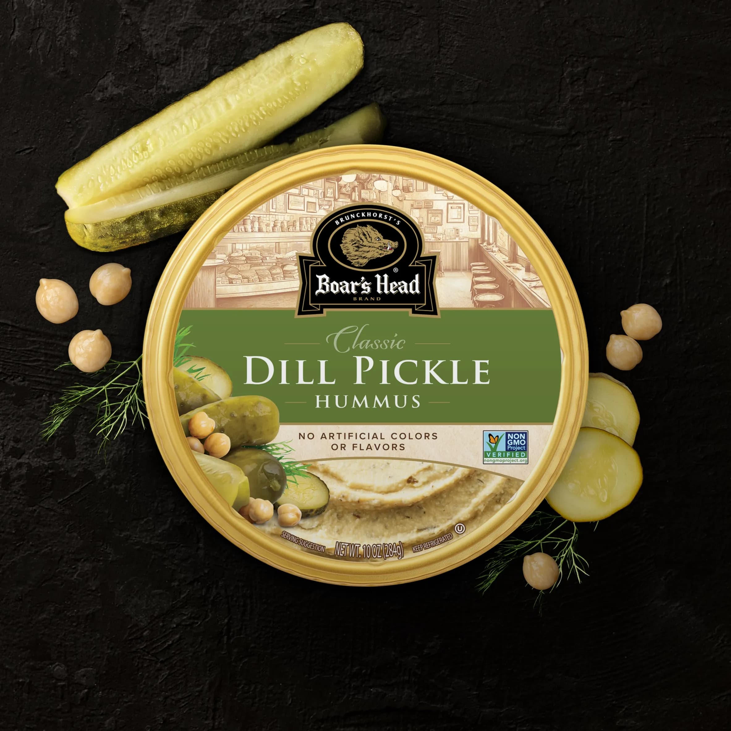 An aerial view of the Boar's Head® Dill Pickle Hummus that sits on a graphite colored background, surrounded by dill pickle slices, whole dill pickles, chickpeas, and dill garnish.