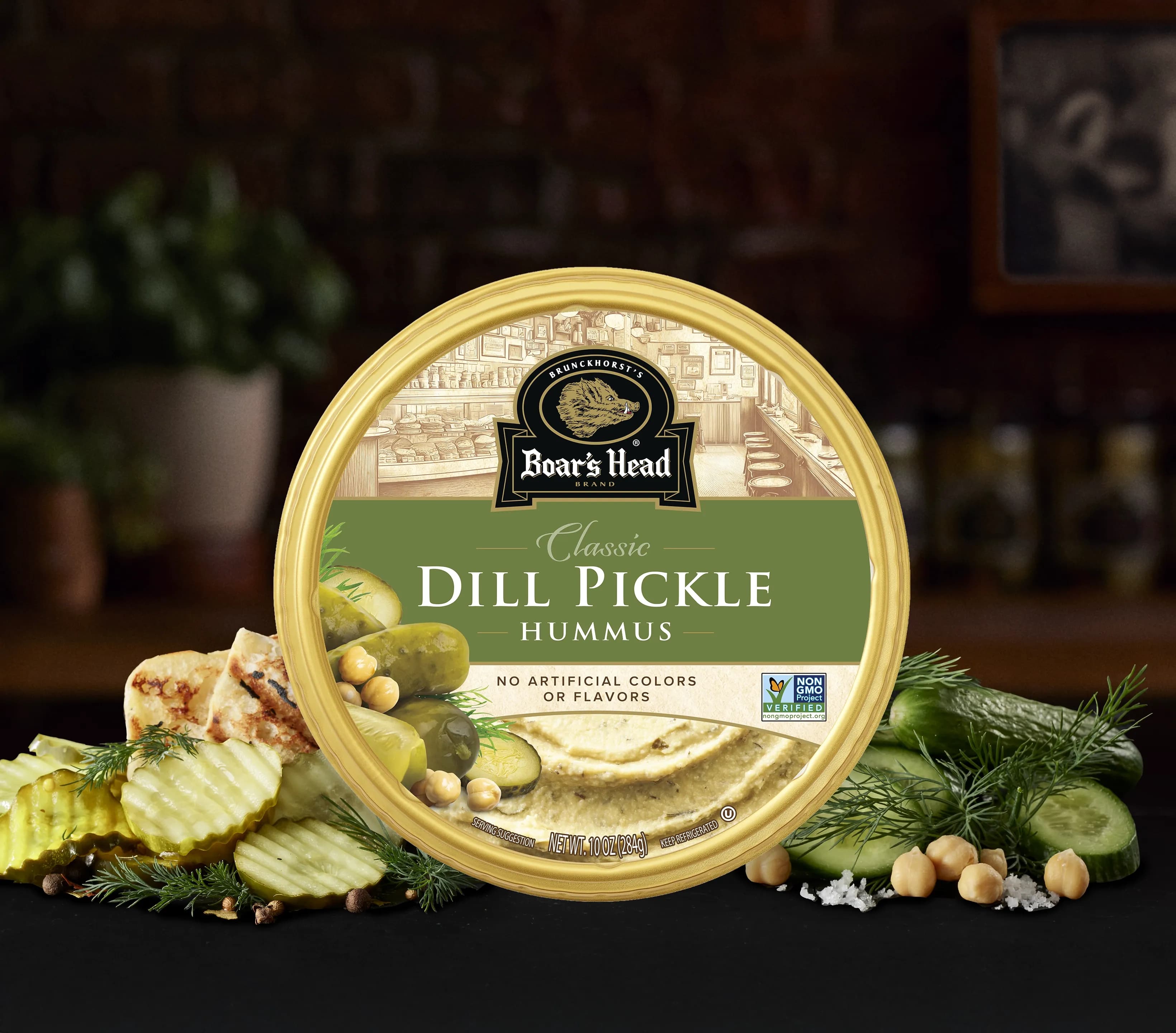The Boar's Head® Dill Pickle Hummus sits on a counter top, surrounded by dill pickle slices, whole dill pickles, chickpeas, and dill garnish, all in front of blurred deli scene in the background.