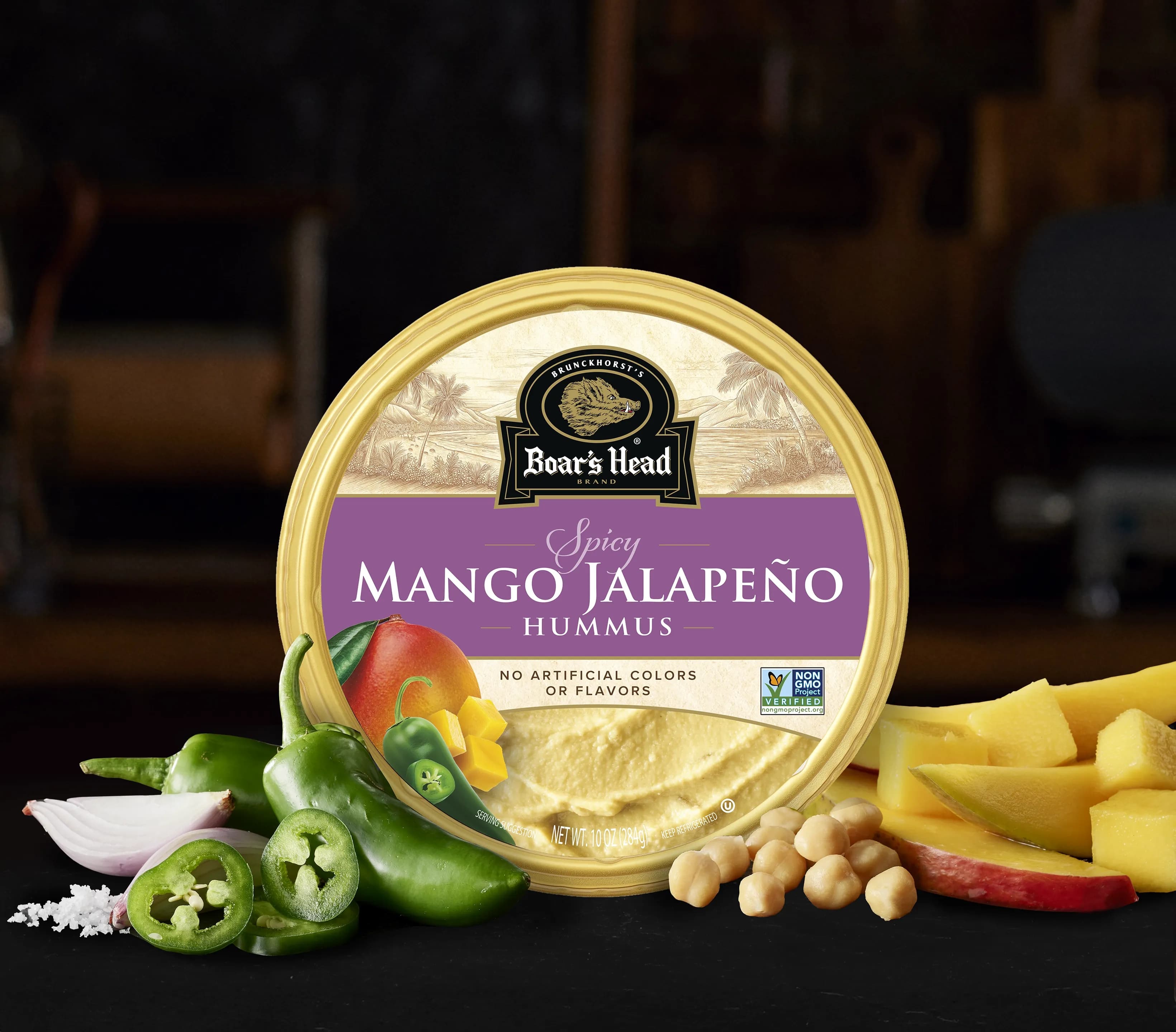 The Boar's Head® Mango Jalapeño Hummus sits on a counter top, surrounded by chickpeas, mango slices, green peppers, and salt, all in front of blurred deli scene in the background.