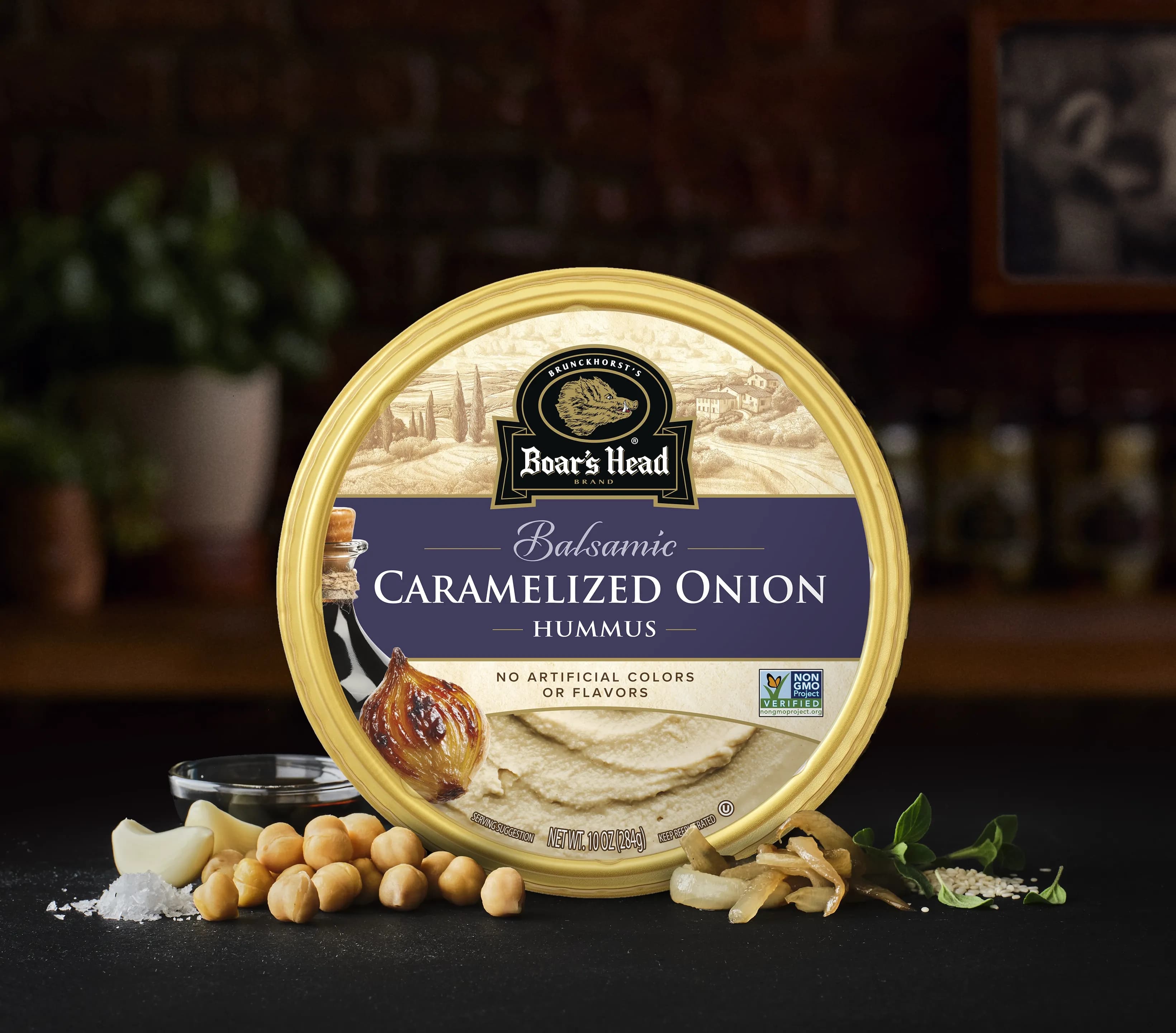 The Boar's Head® Caramelized Onion Hummus sits on a counter top, surrounded by chickpeas, a bowl of balsamic vinegar, caramelized onions, and green garnish, all in front of blurred deli scene in the background.