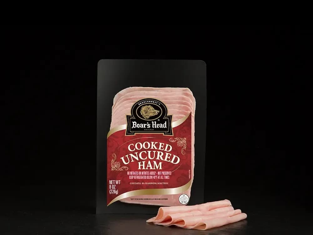A package of Boar's Head® Cooked Uncured Ham sits on a counter surface, with several slices of the product sitting in front of it, both in front of a black background.