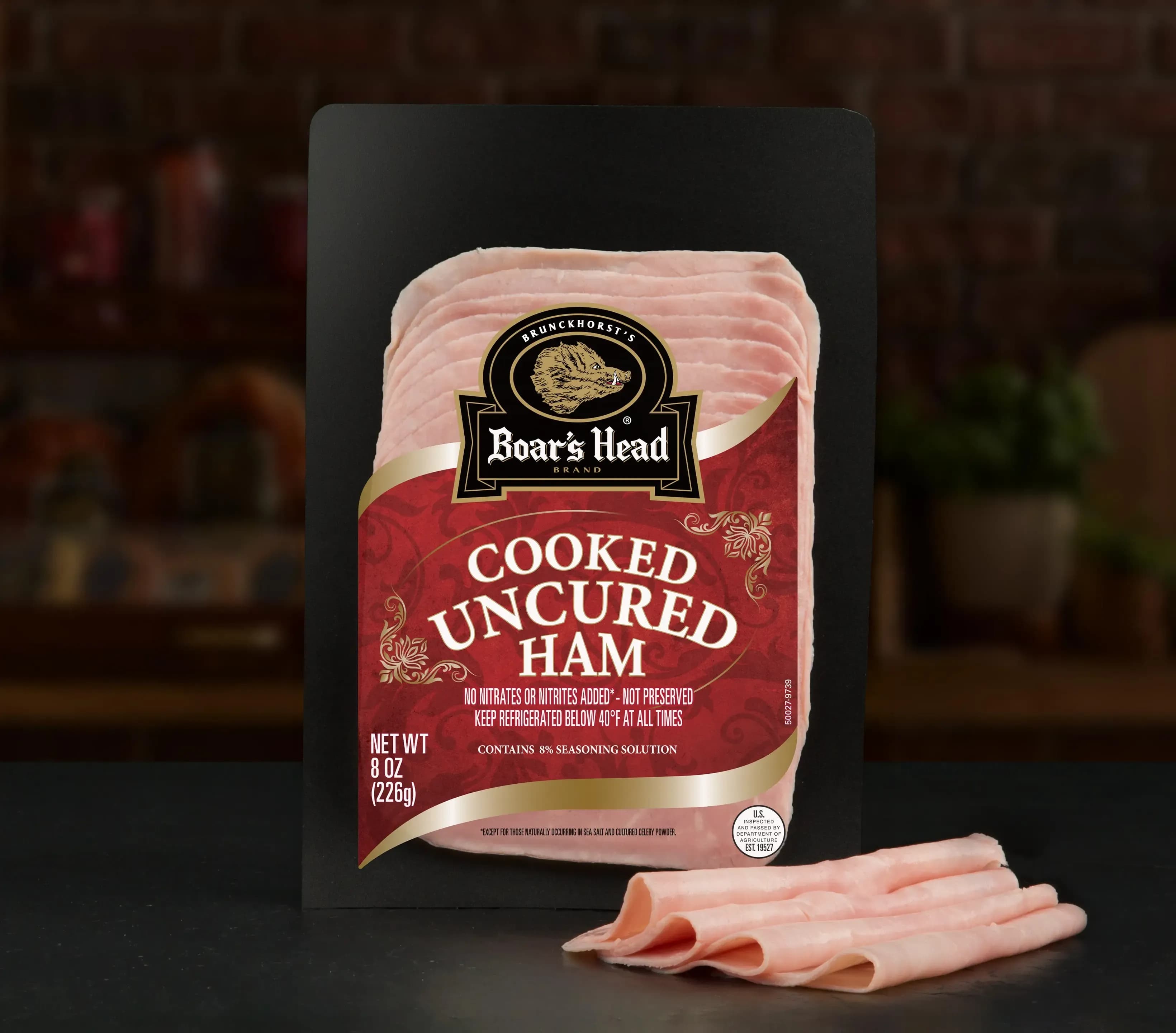 A package of Boar's Head® Cooked Uncured Ham sits on a counter surface, with several slices of the product sitting in front of it, in front of blurred deli scene in the background.
