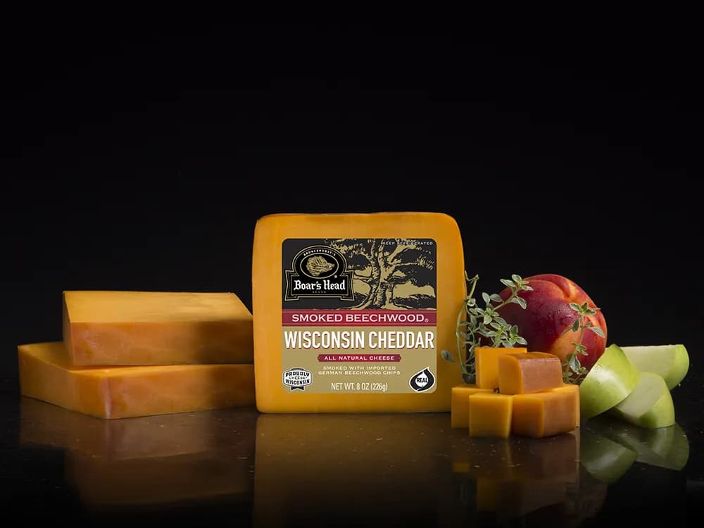 The Boar's Head® Smoked Beechwood® Wisconsin Cheddar Cheese sits on a counter top, surrounded by a plum, green apple slices, greenery, and sliced and cubed product, all in front of black background.