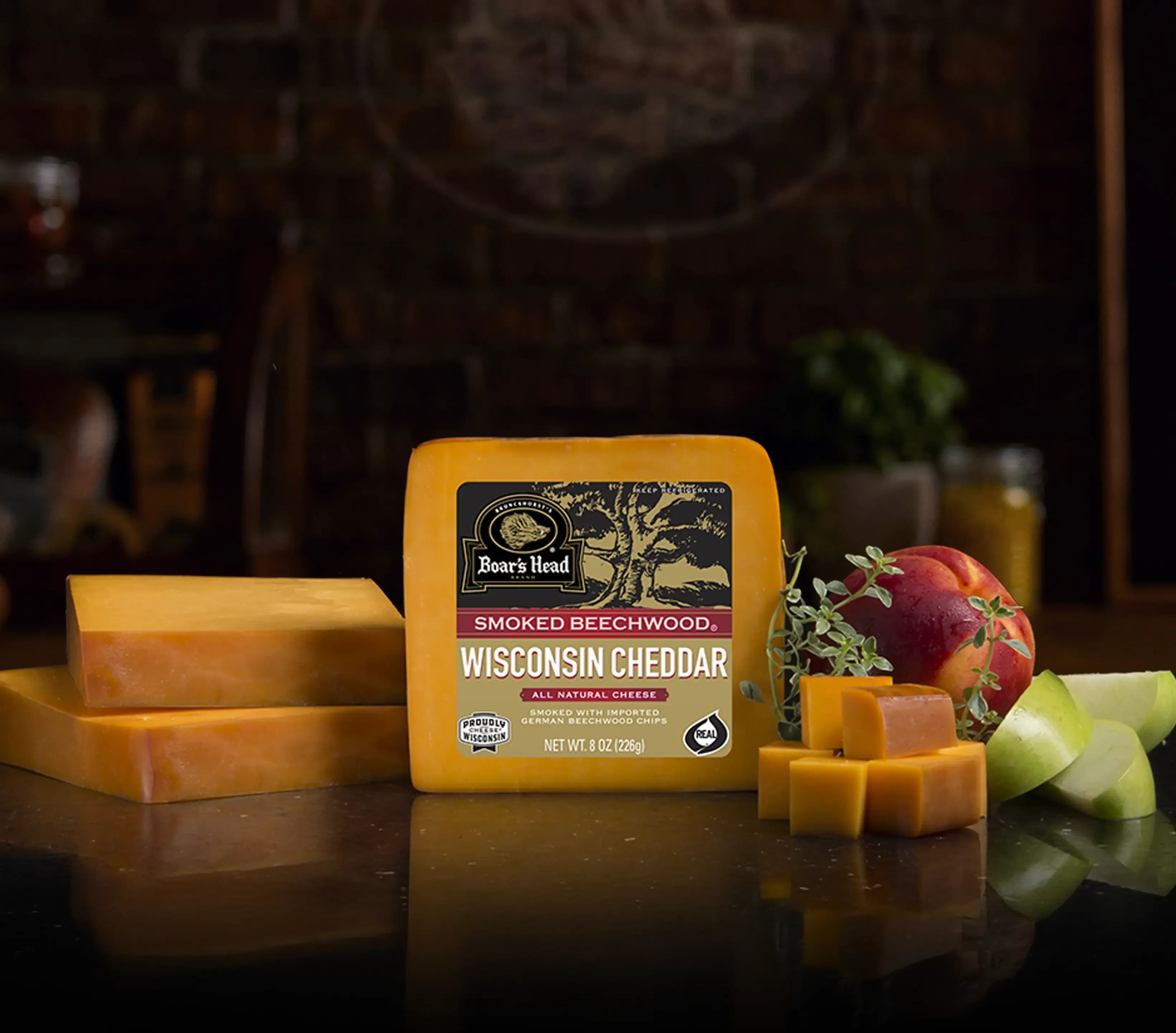 The Boar's Head® Smoked Beechwood® Wisconsin Cheddar Cheese sits on a counter top, surrounded by a plum, green apple slices, greenery, and sliced and cubed product, all in front of blurred deli scene in the background.