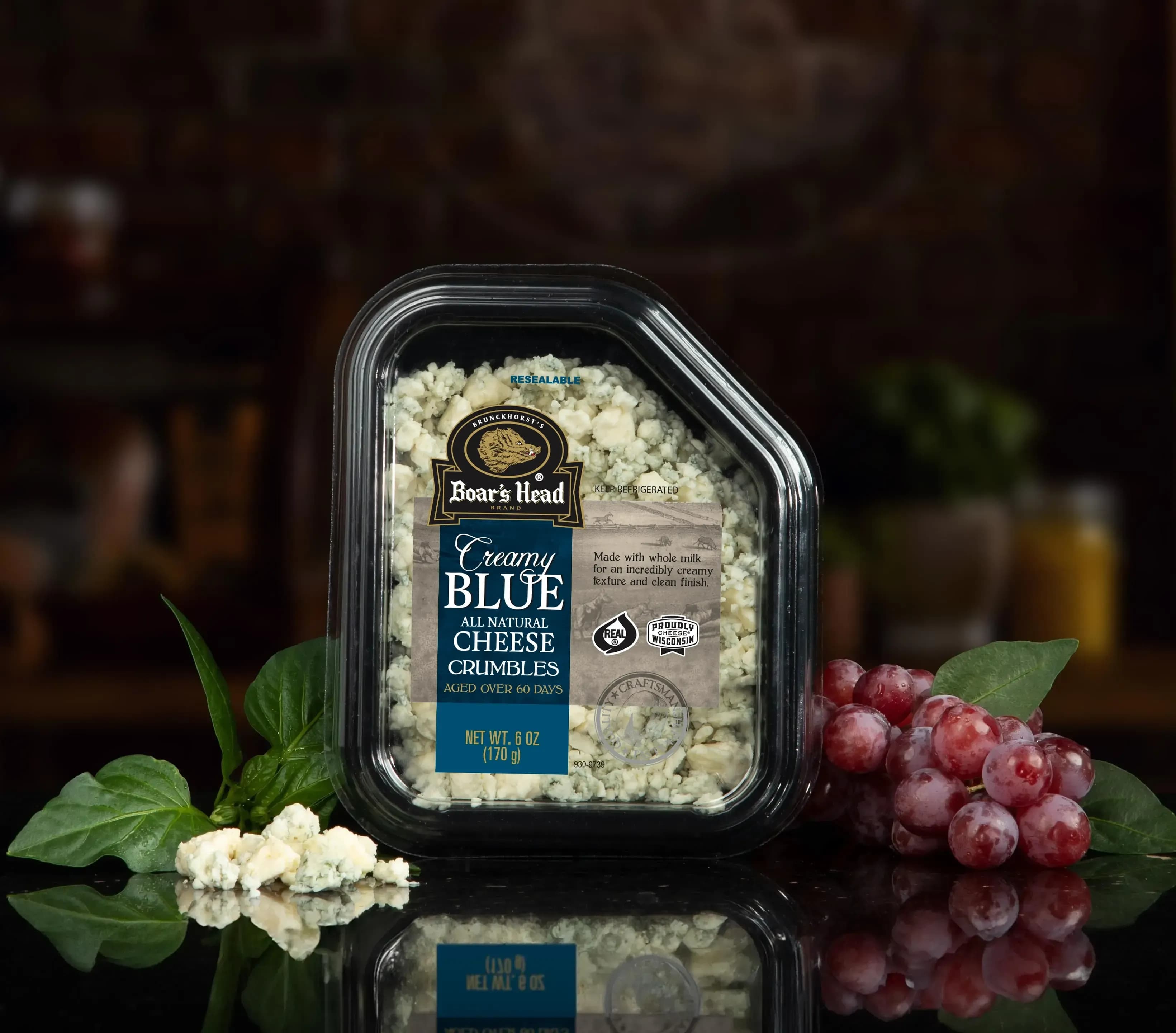 The Boar's Head® Creamy Blue Cheese Crumbles sits on a counter top, surrounded by grapes,, greenery, and crumbled product, all in front of blurred deli scene in the background.