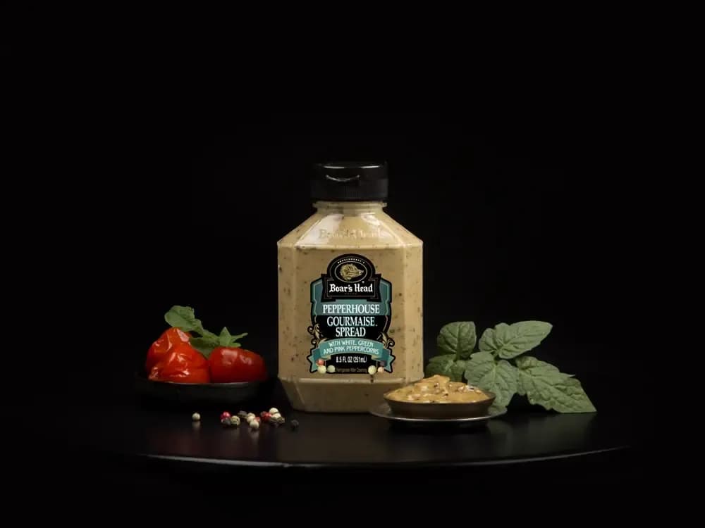 The Boar's Head® Pepperhouse Gourmaise® Spread sits on a counter top, surrounded by roasted tomatos, peppercorns, greenery, and a container of the spread, all in front of black background.