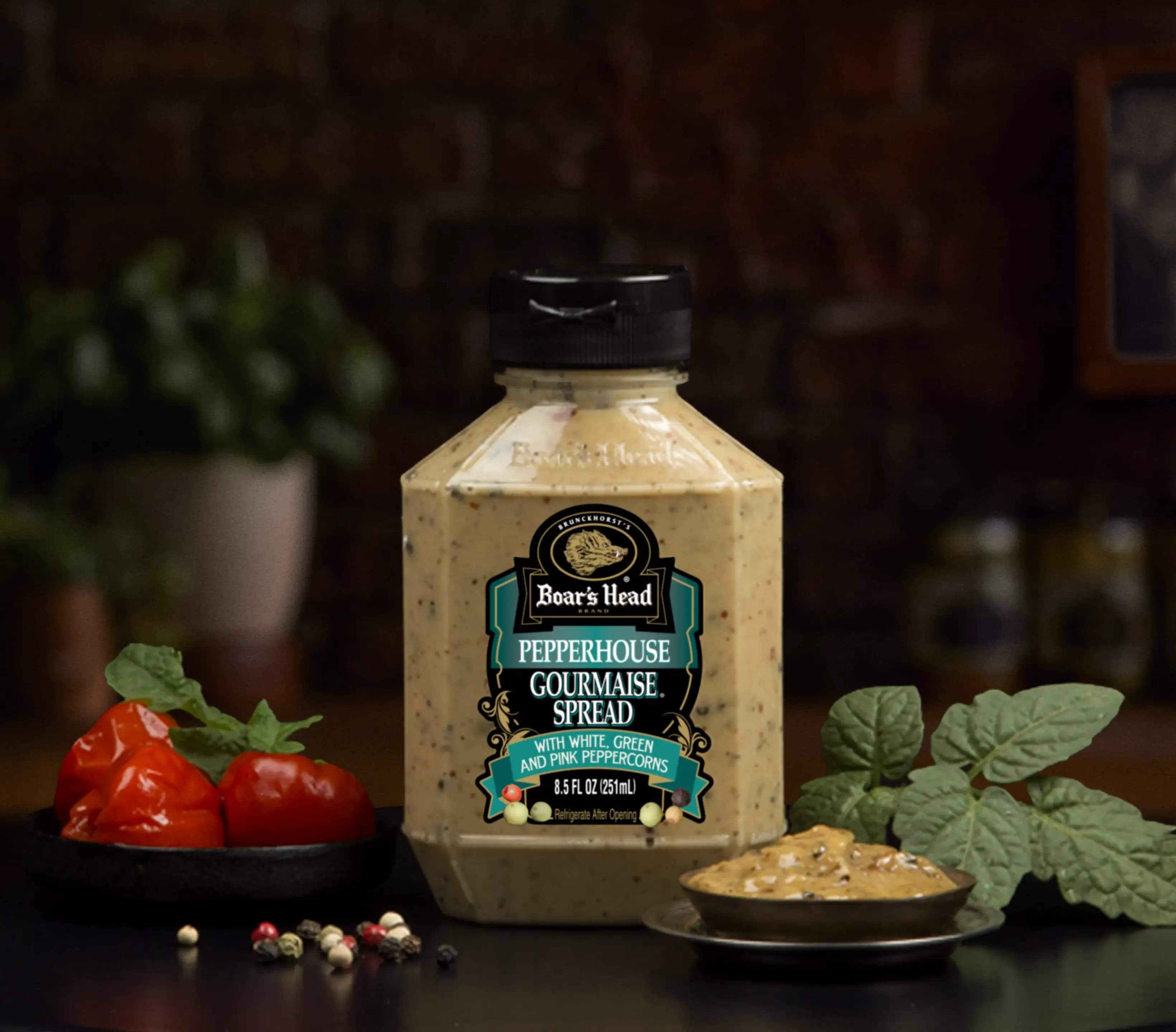 The Boar's Head® Pepperhouse Gourmaise® Spread sits on a counter top, surrounded by roasted tomatos, peppercorns, greenery, and a container of the spread, all in front of black background.