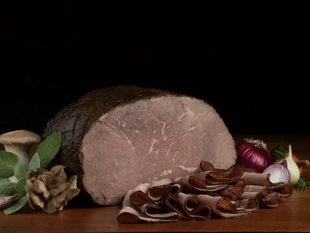 The Boar's Head® Seasoned Filet of Roast Beef - Well Done sits on a counter top, surrounded by mushrooms, red onions, greenery, and sliced product, all in front of black background.