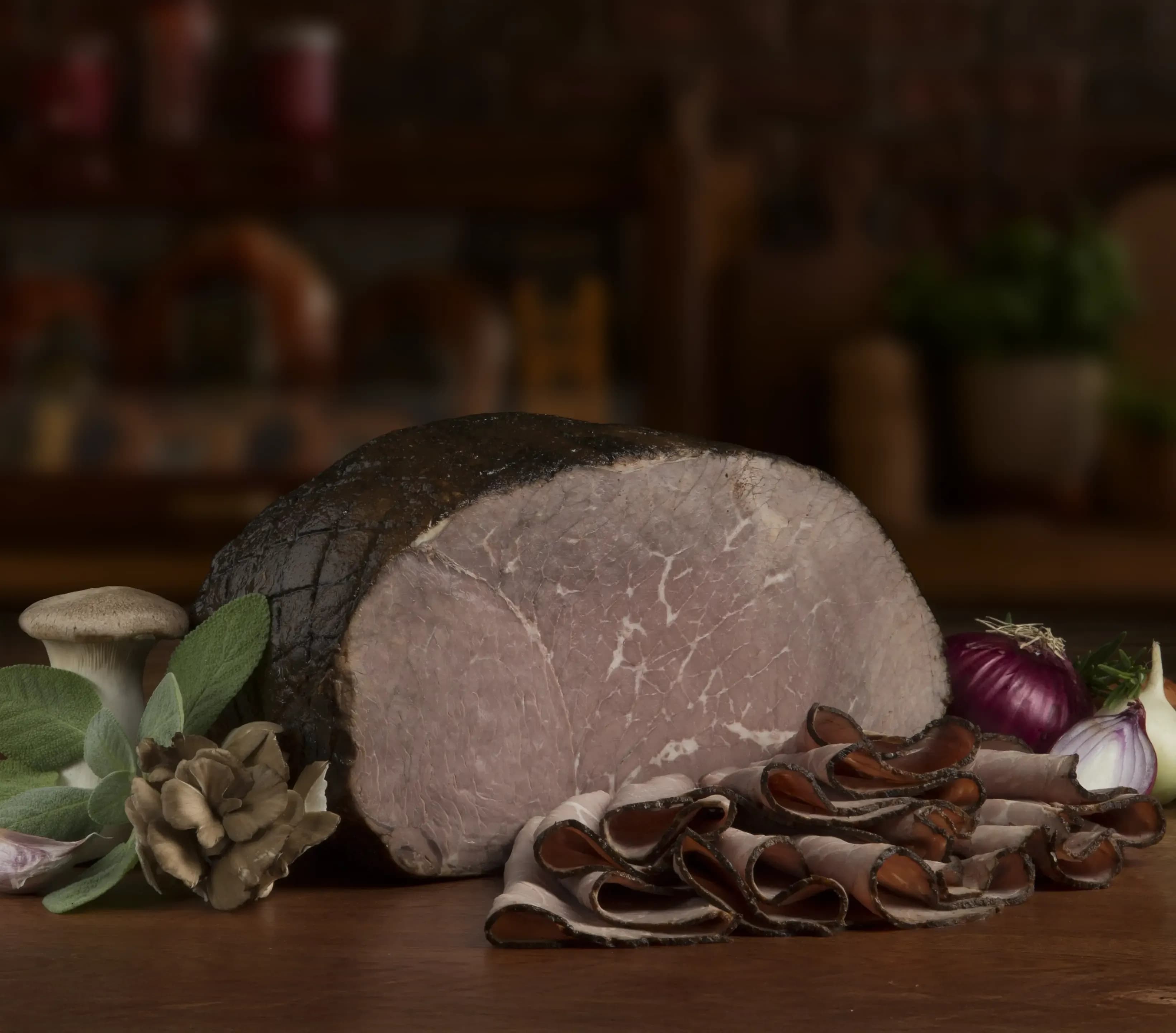 The Boar's Head® Seasoned Filet of Roast Beef - Well Done sits on a counter top, surrounded by mushrooms, red onions, greenery, and sliced product, all in front of blurred deli scene in the background.