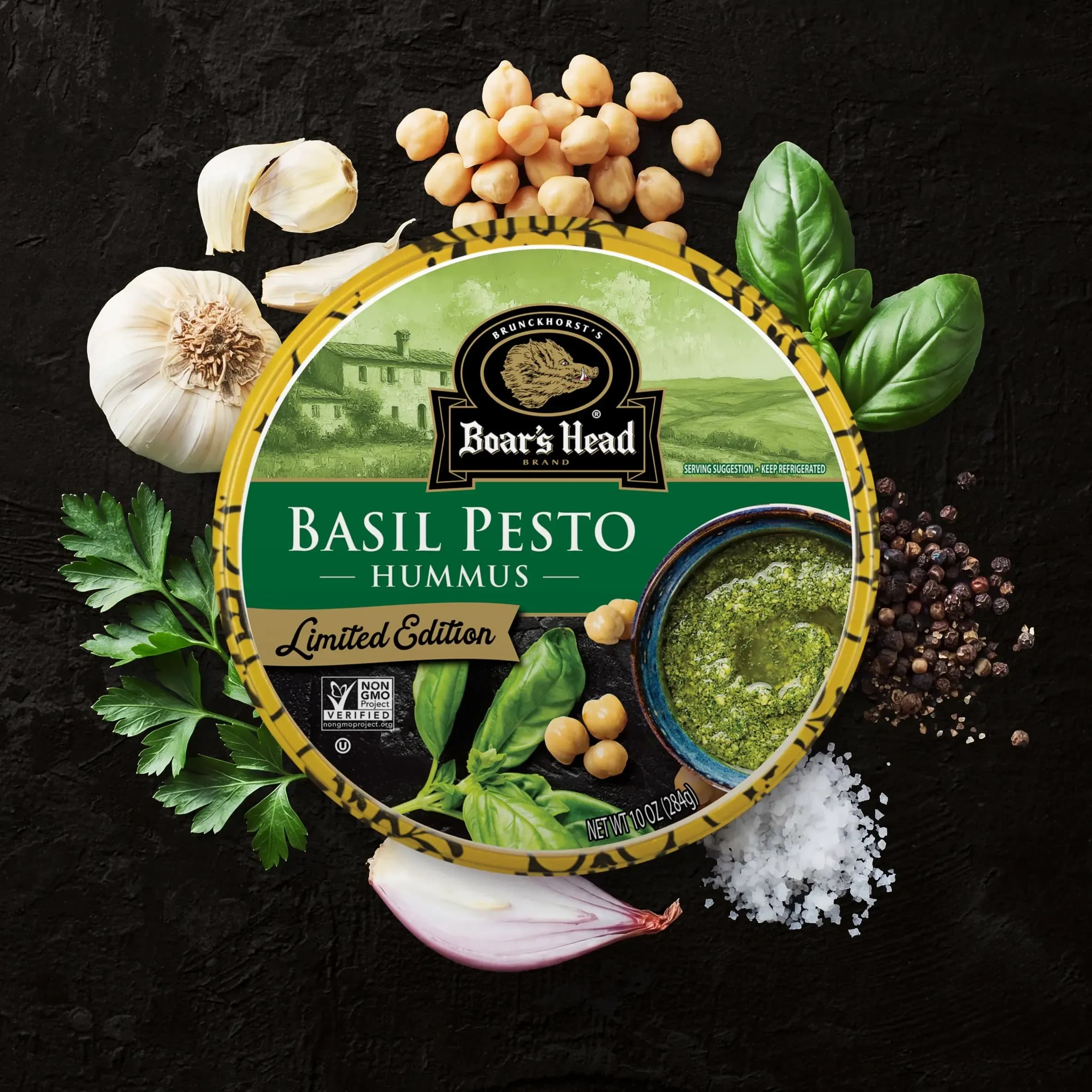An aerial view of the Boar's Head® Basil Pesto Hummus that sits on a graphite colored background, surrounded by basil leaves, green garnish, chickpeas, garlic, salt, and pepper.