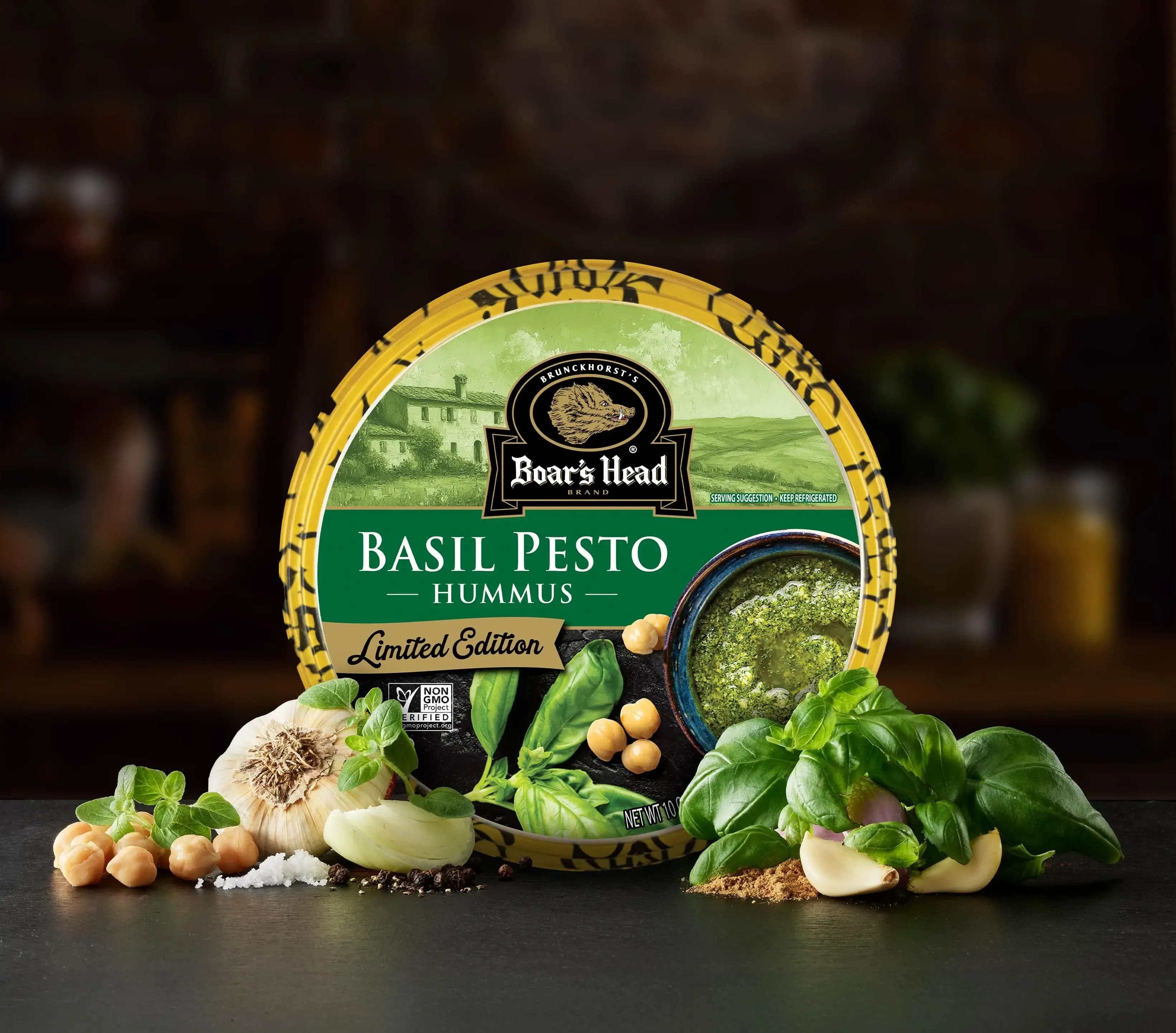 The Boar's Head® Basil Pesto Hummus sits on a counter top, surrounded by basil leaves, green garnish, chickpeas, garlic, salt, and pepper.