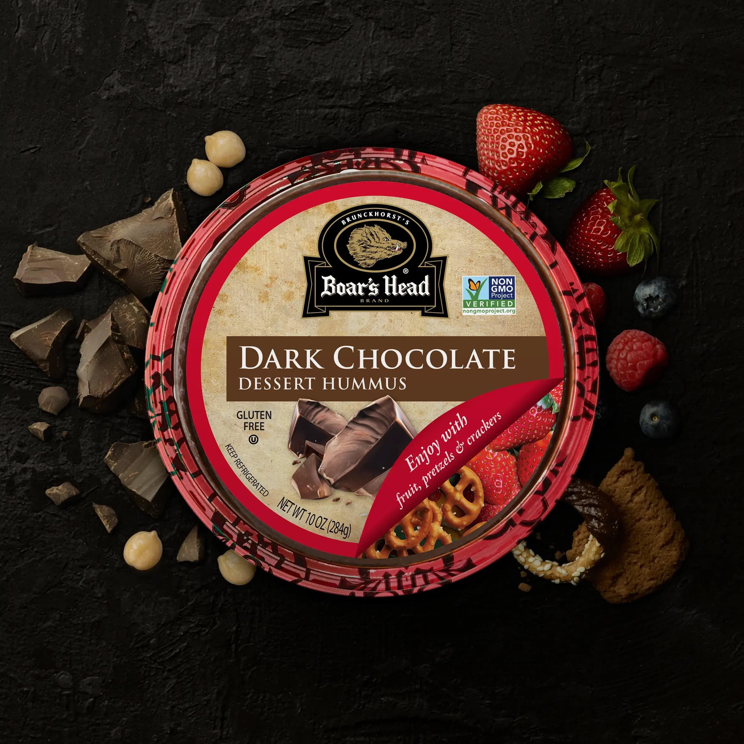 An aerial view of the Boar's Head® Dark Chocolate Hummus that sits on a graphite colored background, surrounded by dark chocolate pieces, raspberries, strawberries, pineapple slices, and chickpeas.